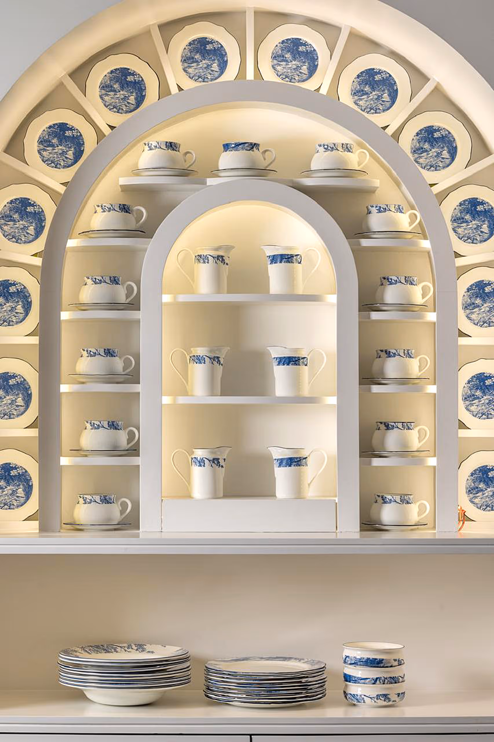 Crockery display design by KRT Design Studio, featuring blue-and-white illustrated porcelain plates and cups arranged within a softly illuminated arched cabinet, reflecting heritage-inspired illustration, refined spatial composition, and a curated luxury dining experience.