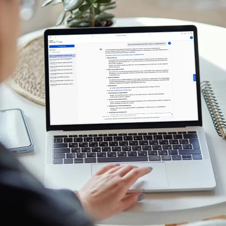 Eine Person arbeitet an einem Laptop mit geöffnetem Dokument, umgeben von Smartphone, Notizbuch und Pflanze auf einem Tisch und nutzt den Haufe HR Assistant.