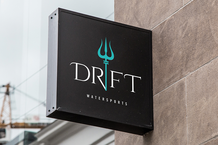 Black rectangular sign mounted on a wall displaying the text 'DRIFT' with a turquoise trident icon integrated into the letter 'I' and the word 'WATERSPORTS' below in smaller letters.