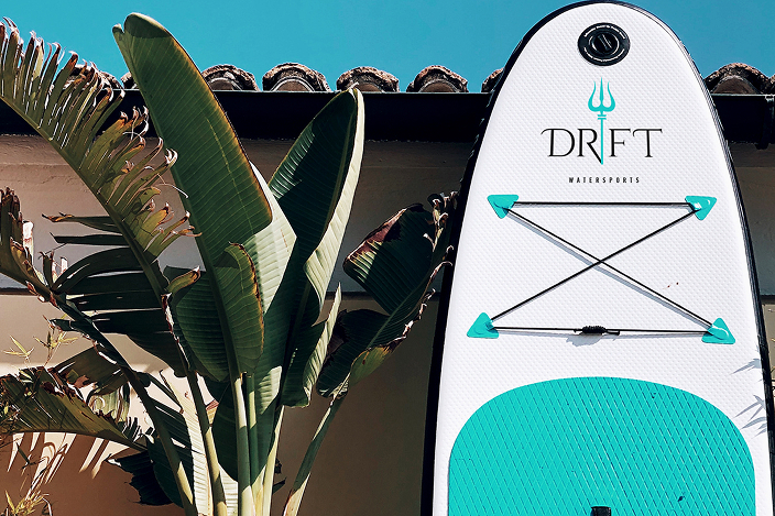 Partially visible white and turquoise stand-up paddleboard with DRIFT Watersports logo leaning against a wall next to tropical green leaves and blue sky.