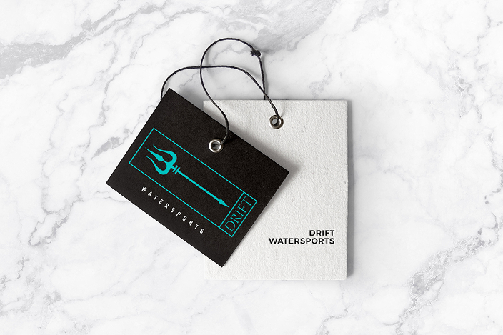 Two product tags on a marble surface; one black with a turquoise trident logo and 'DRIFT WATERSPORTS' text, and one white with 'DRIFT WATERSPORTS' text in black.