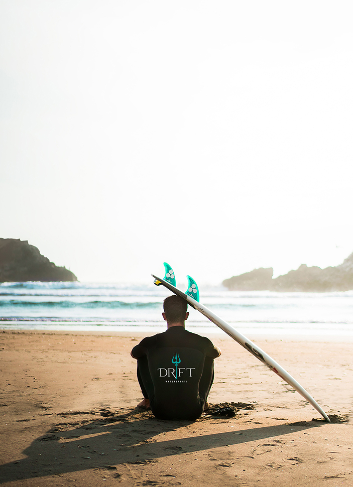 Surfer sitting on sandy beach with surfboard resting on his shoulder, facing the ocean waves at sunrise or sunset.