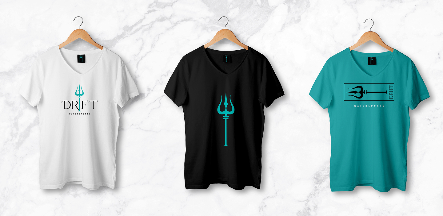 Three V-neck T-shirts on wooden hangers against a marble background: white with turquoise trident and DRIFT logo, black with large turquoise trident, and teal with black trident in a rectangular frame and DRIFT logo.