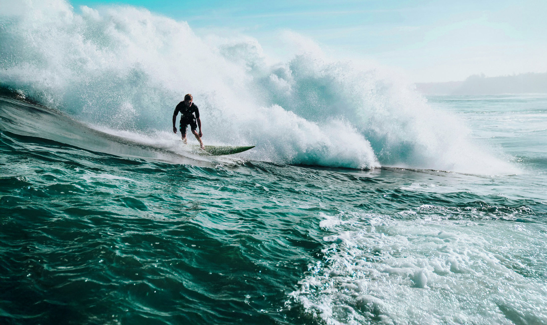 Surfer riding a large wave on turquoise ocean water under a clear blue sky.