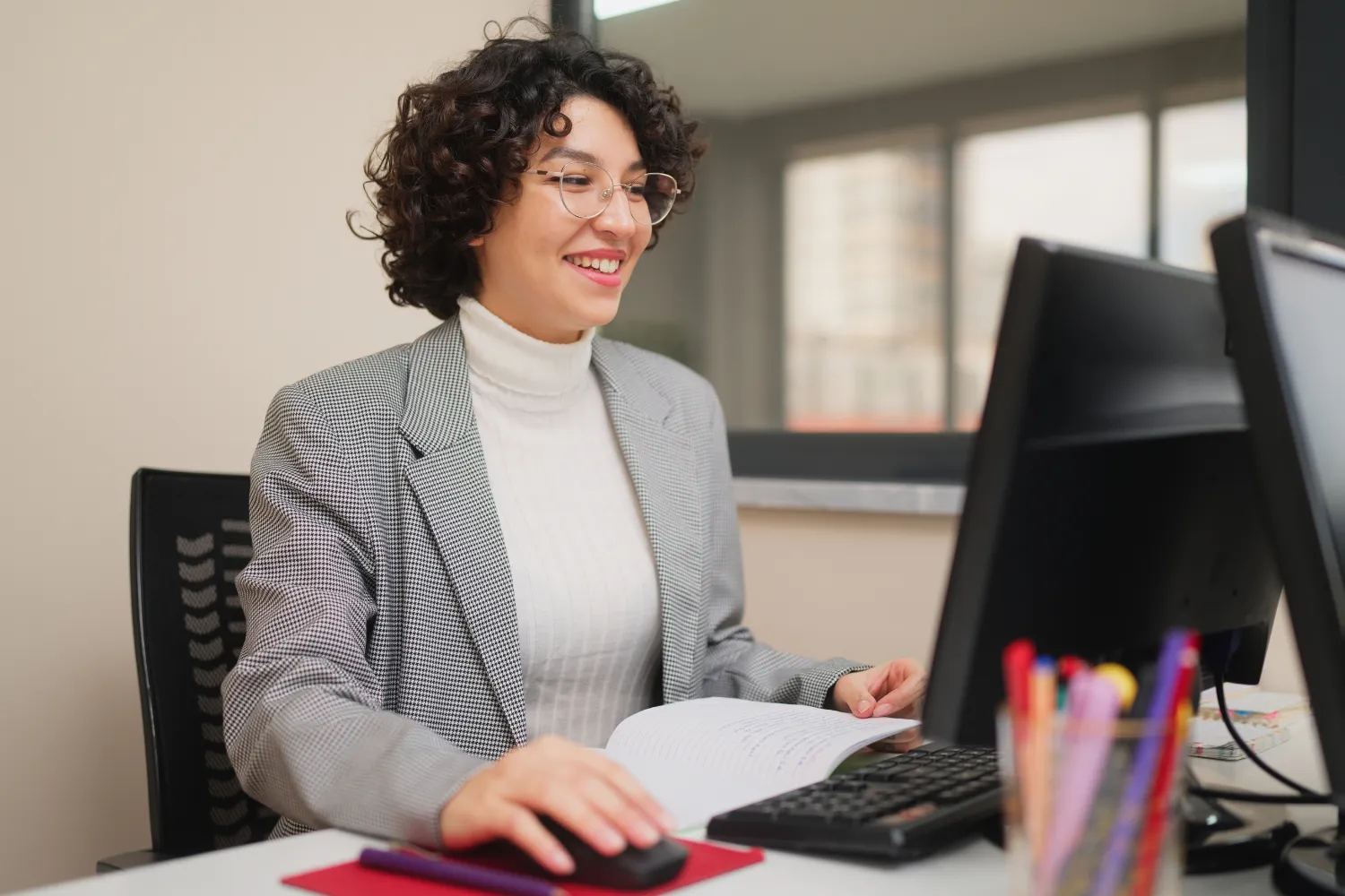 Smiling woman in glasses and a checked blazer working on a computer while holding an open notebook.