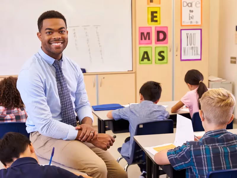 A man wearing a blue shirt and tie is sitting in front of a classroom.