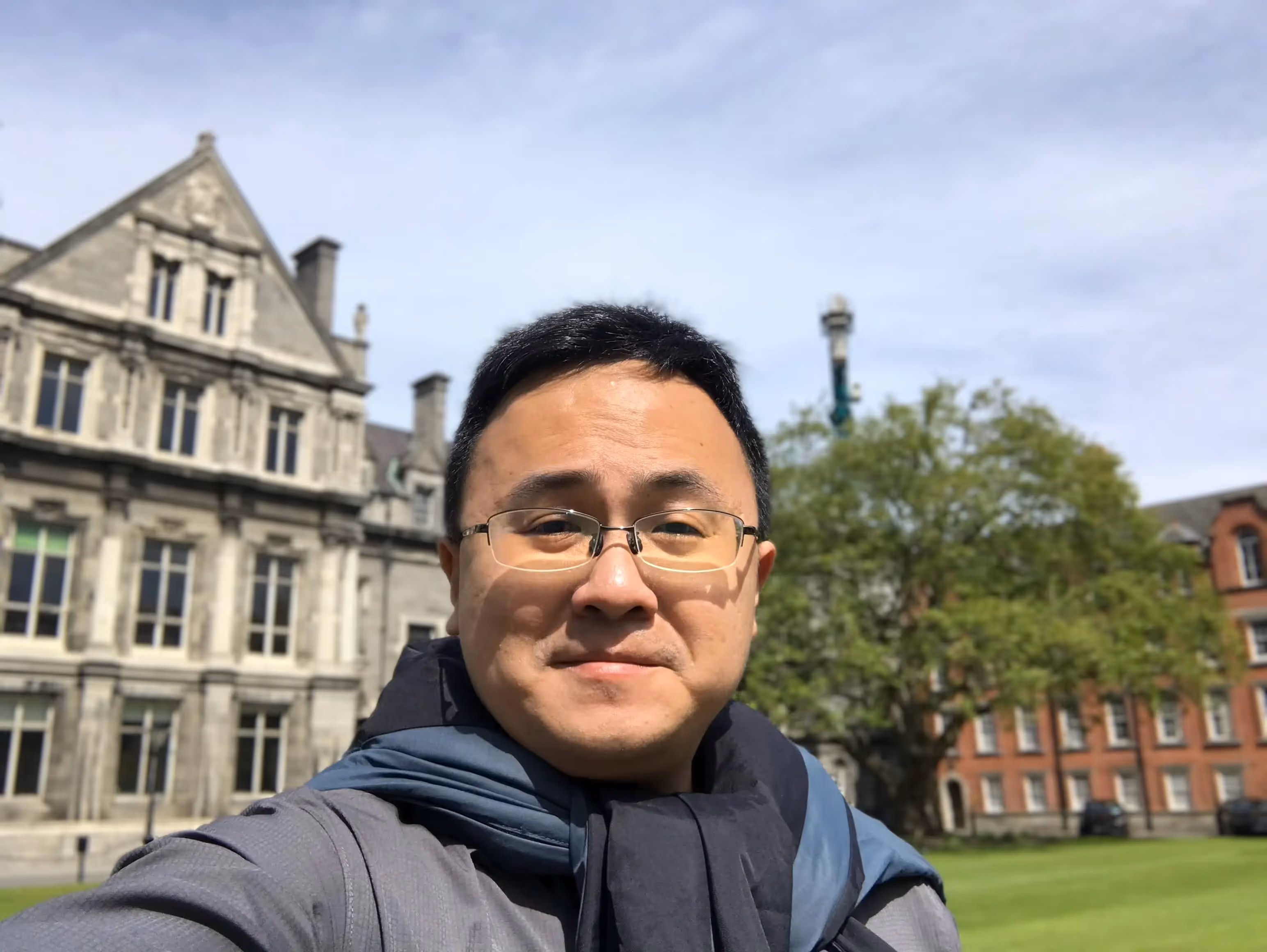 A man wearing glasses and a blue scarf stands in front of a building. - Assistments - Formative Assessment Solutions