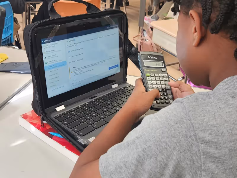 A young girl is using a laptop and a calculator.. - ASSISTments Images