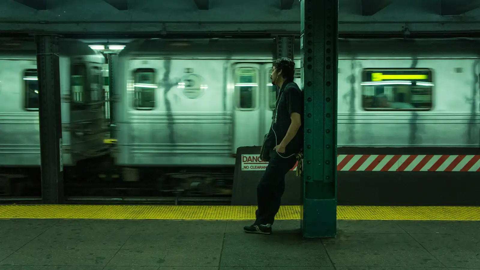 A man leaning against a metal pillar in the subway, listening to music.
