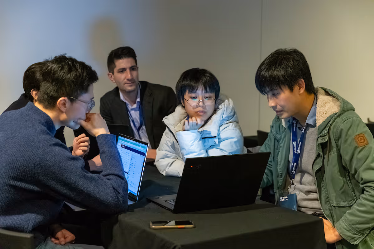 Five people sit around a table, focused on two laptops during a group discussion.