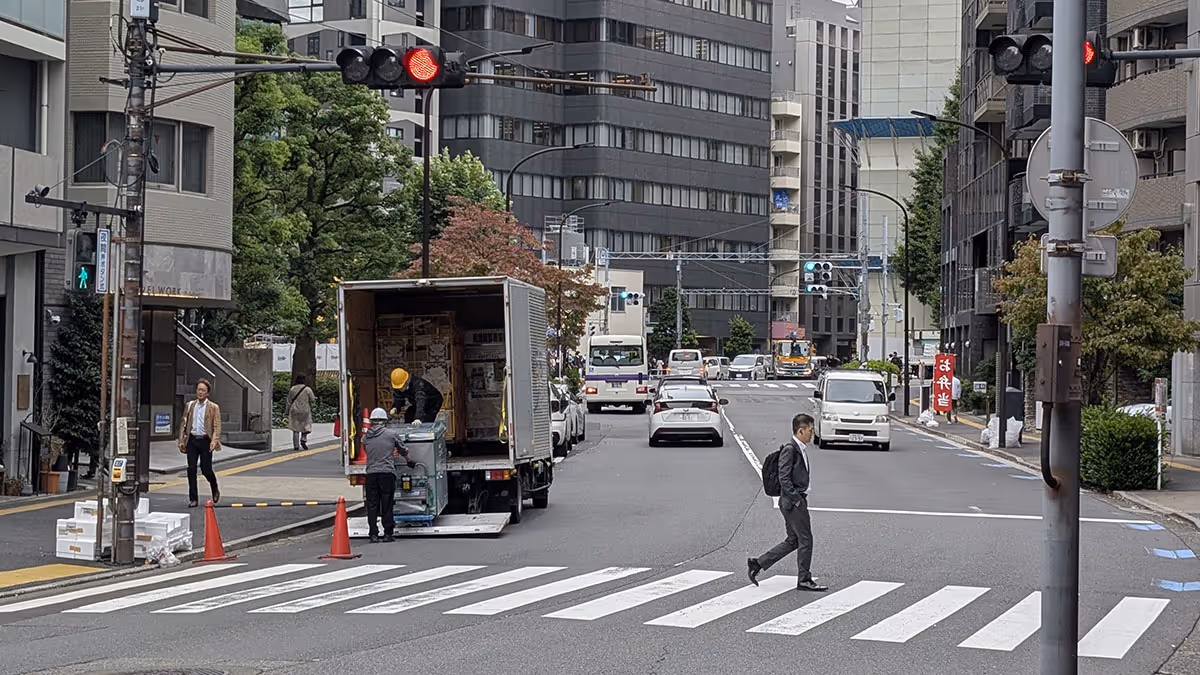 Two workers unloading boxes from a truck parked on a busy Tokyo street while pedestrians cross the street at a crosswalk.