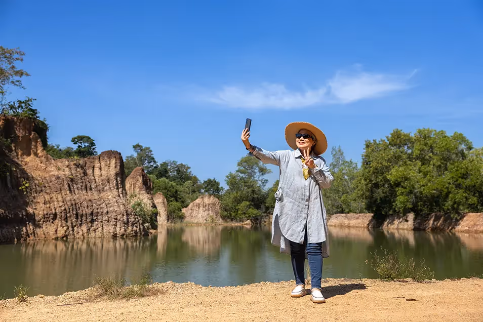 A woman wearing a sunhat and sunglasses takes a selfie by a scenic lake surrounded by rocky cliffs and lush greenery under a clear blue sky.