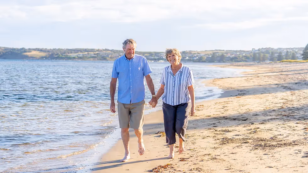 Smiling older Australian couple walking barefoot along a sunny beach, enjoying a relaxing and safe travel experience.