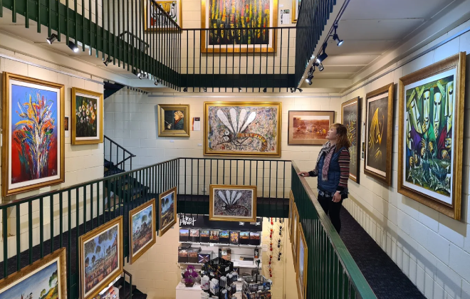 Interior of Pro Hart Gallery with multiple levels displaying colourful framed paintings and a visitor observing the artworks.