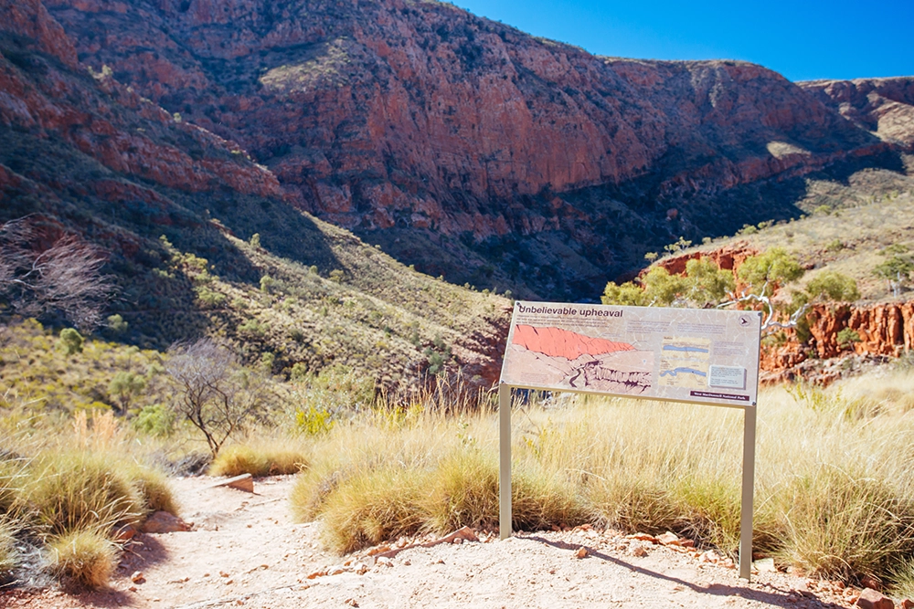 Interpretive sign overlooking Ormiston Gorge in the West MacDonnell Ranges, with red rock cliffs and dry grasses under a clear blue sky.