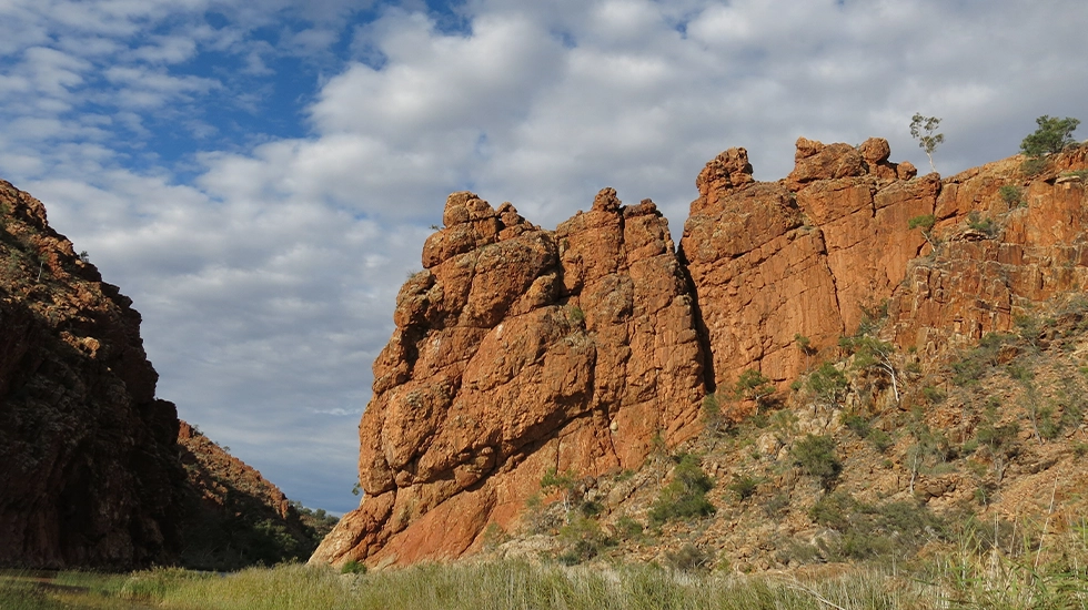 Red rock cliffs and rugged gorge at Ellery Creek Big Hole under a blue sky with scattered clouds.