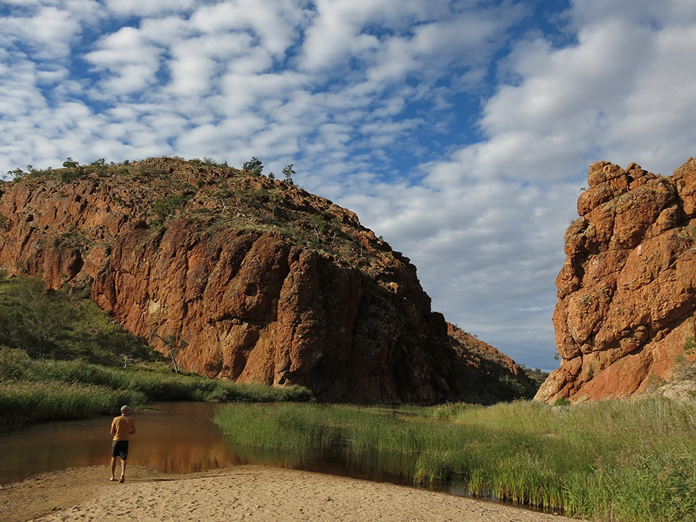 Person standing by Ellery Creek Big Hole with red rock cliffs, sandy bank and calm water under a partly cloudy sky.