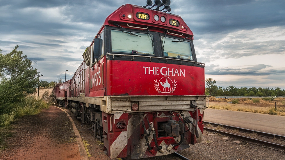 The Ghan locomotive at a rural station under a dramatic, cloudy sky in the Australian outback.