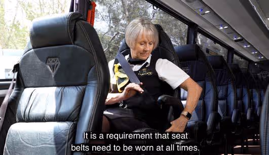 Tour host demonstrating how to fasten a seatbelt, with on-screen text stating seat belts must be worn at all times
