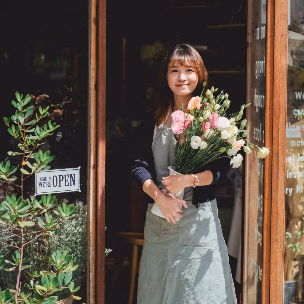 A woman smiling holding flowers in front of her shop.
