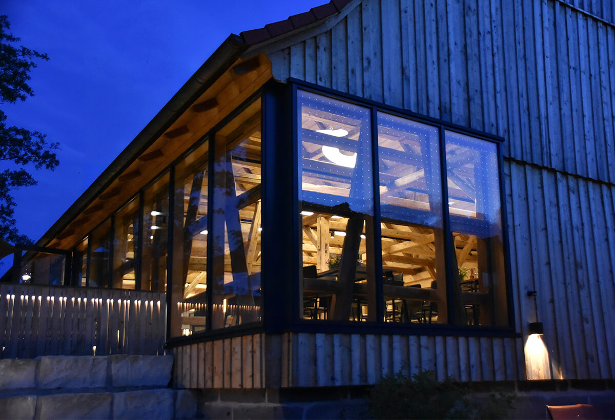 A dark, atmospheric exterior view of the Heinersreuther Hof, featuring an illuminated, glass-enclosed addition with exposed wooden beams.