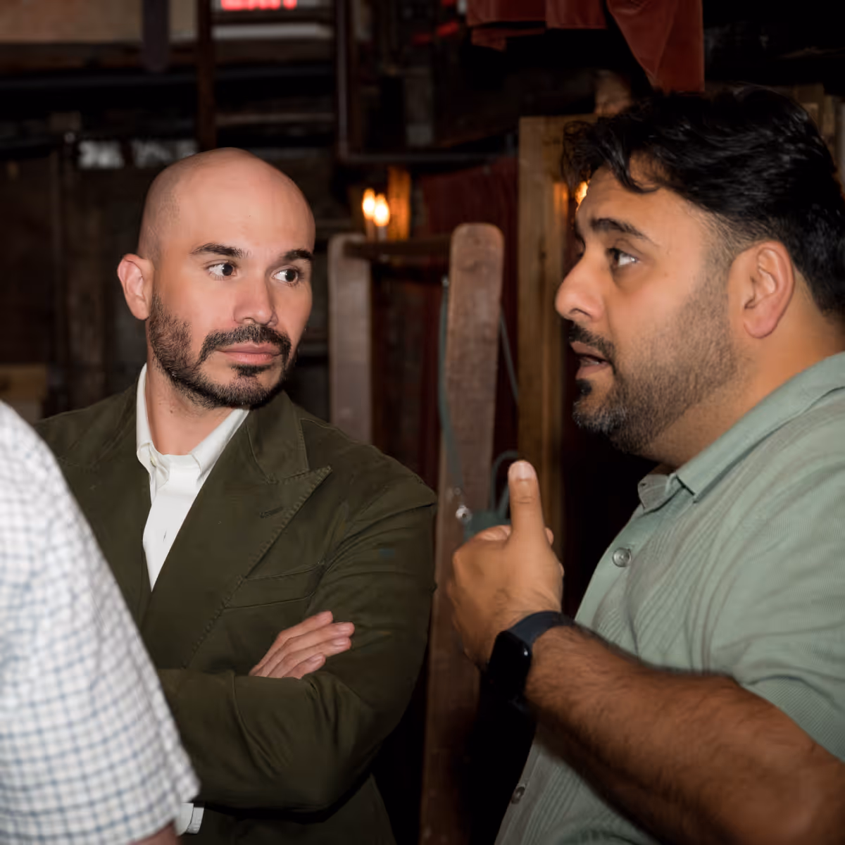 Two men engaged in conversation in a dimly lit indoor setting, one with a bald head and green jacket, the other with dark hair and a light green shirt.