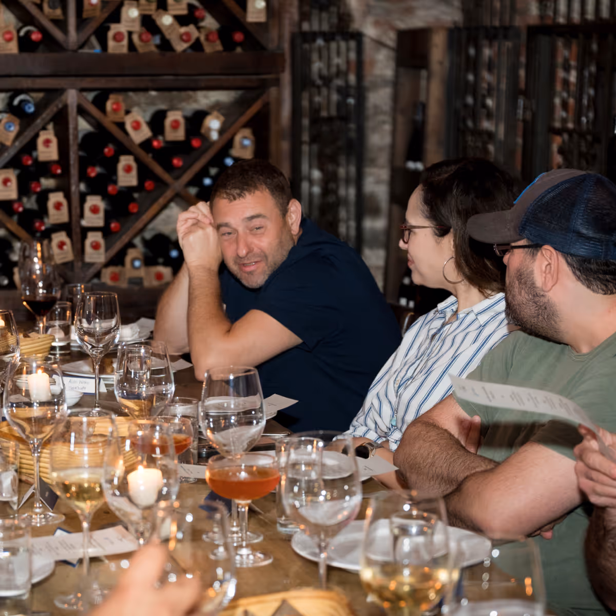 Group of people seated at a table with wine glasses and candles, engaged in conversation in a cozy wine cellar.