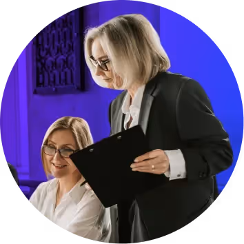 Two professional women in business attire reviewing a clipboard together in an office setting with blue lighting.