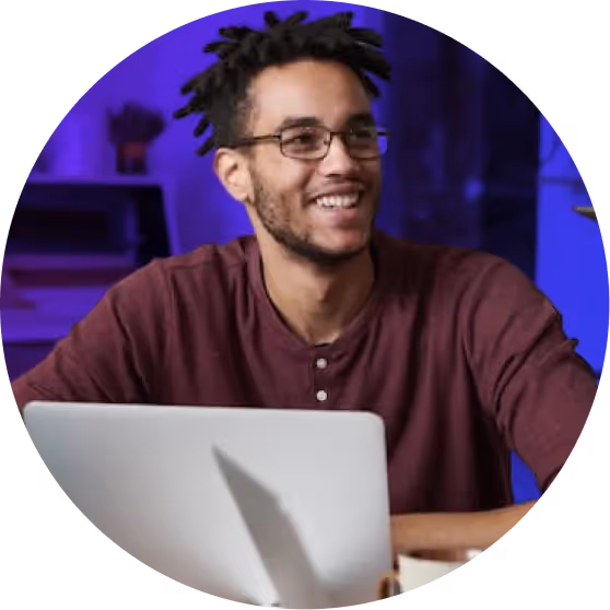 Smiling man with glasses and dreadlocks sitting behind a laptop in a room with purple lighting.