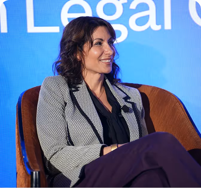 Smiling woman with wavy hair wearing a patterned blazer and black blouse sitting in a brown chair against a blue background.