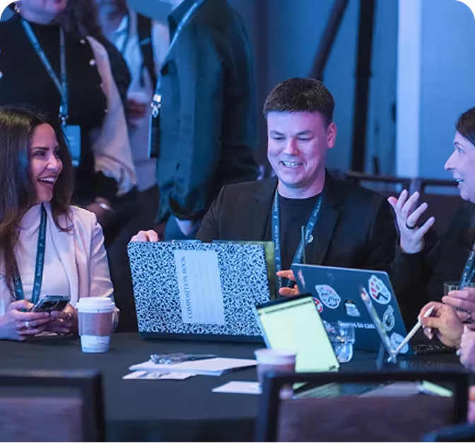 Three professionals smiling and working together at a table with laptops and coffee cups.