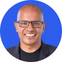 Smiling bald man with glasses wearing a black jacket against a bright blue background.