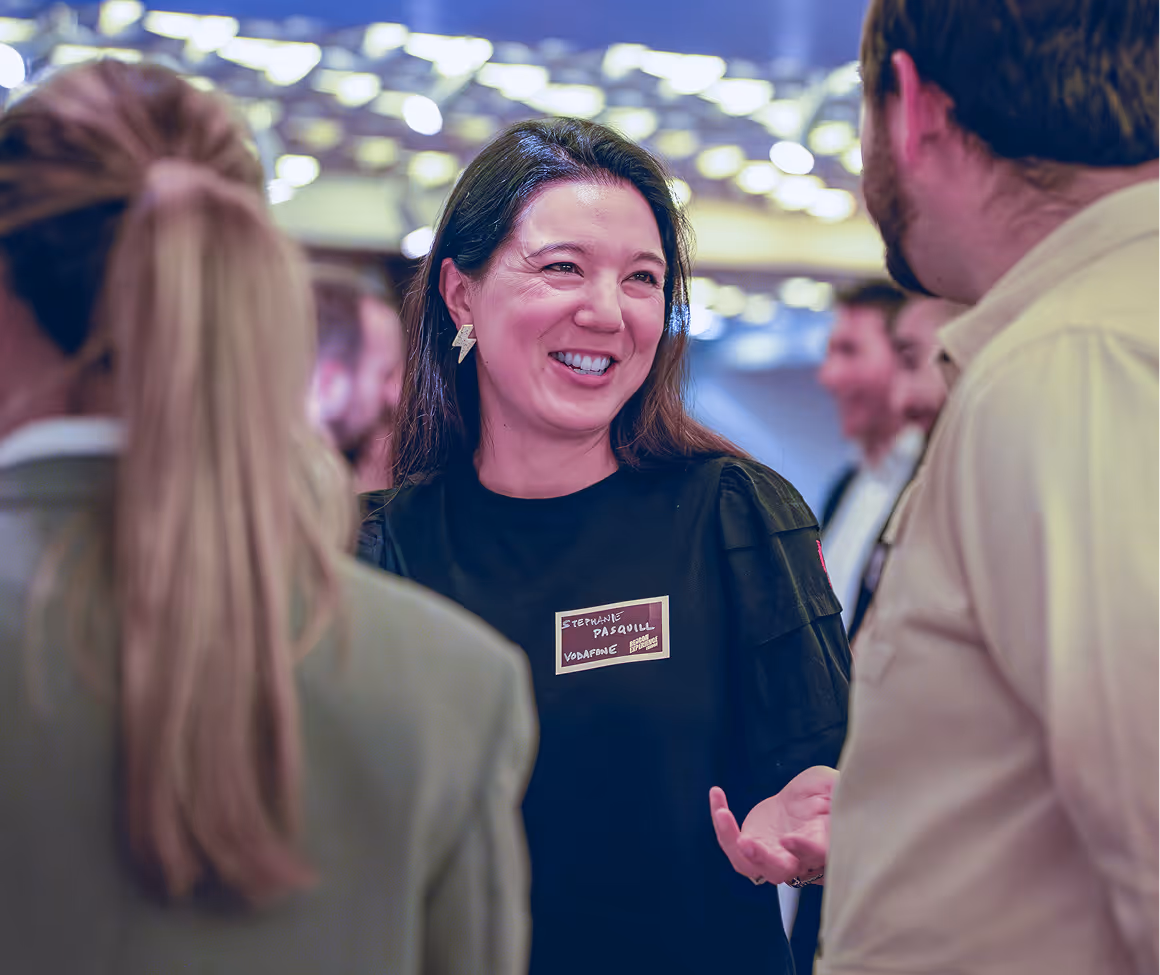 Smiling woman wearing lightning bolt earrings and a nametag talking to two people at a social event with blurred lights in the background.