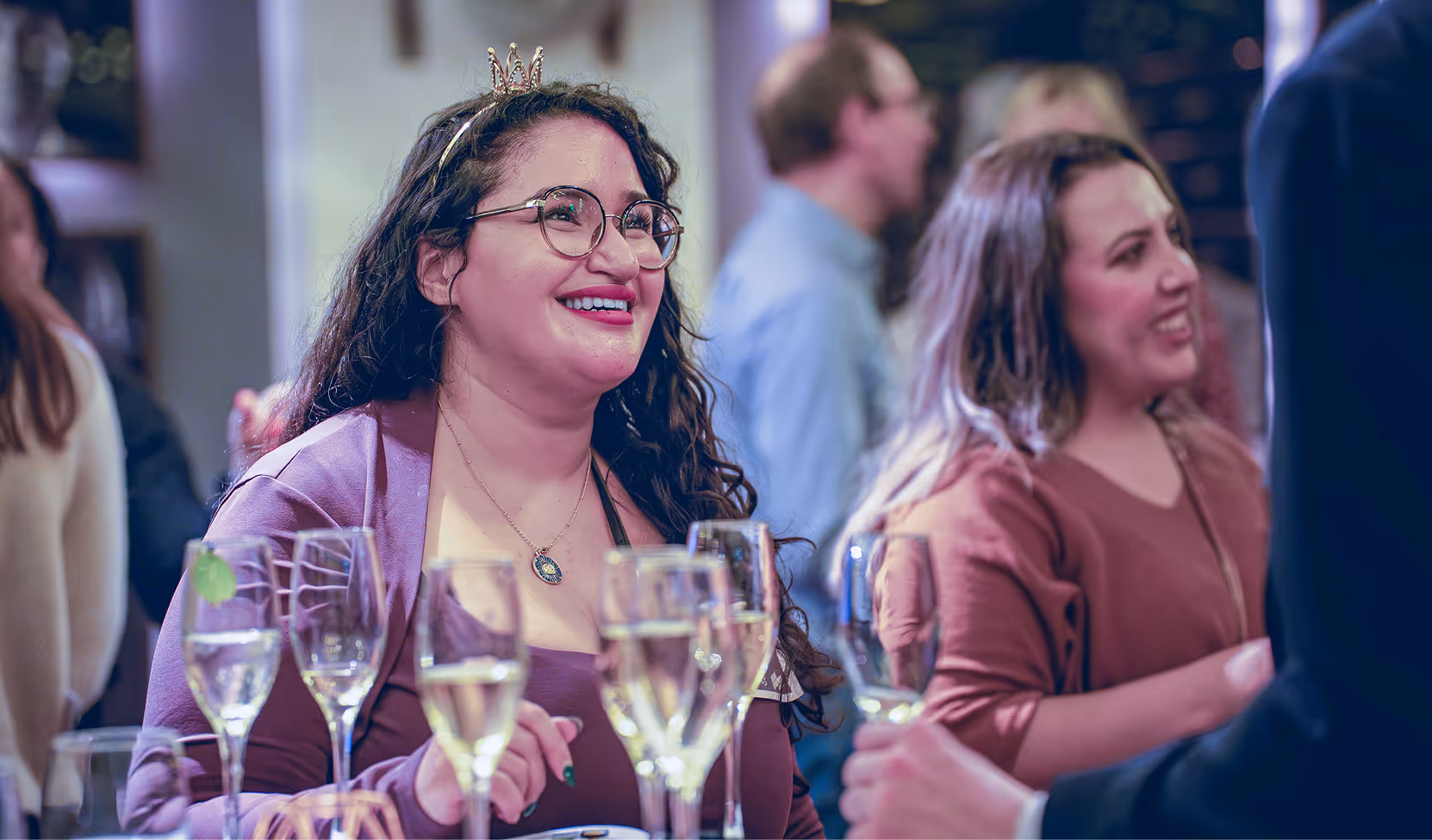 Smiling woman wearing glasses and a small crown headband at a social gathering with glasses of champagne in front of her.