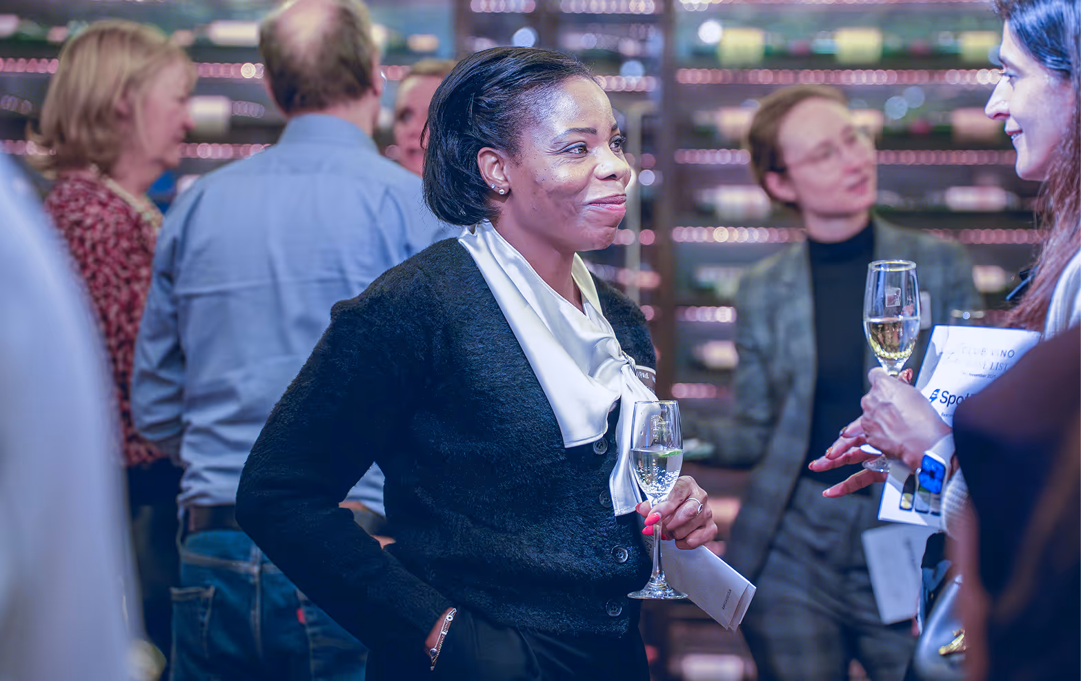 Woman in black sweater and white scarf holding a glass of white wine and talking to another woman at a social event.