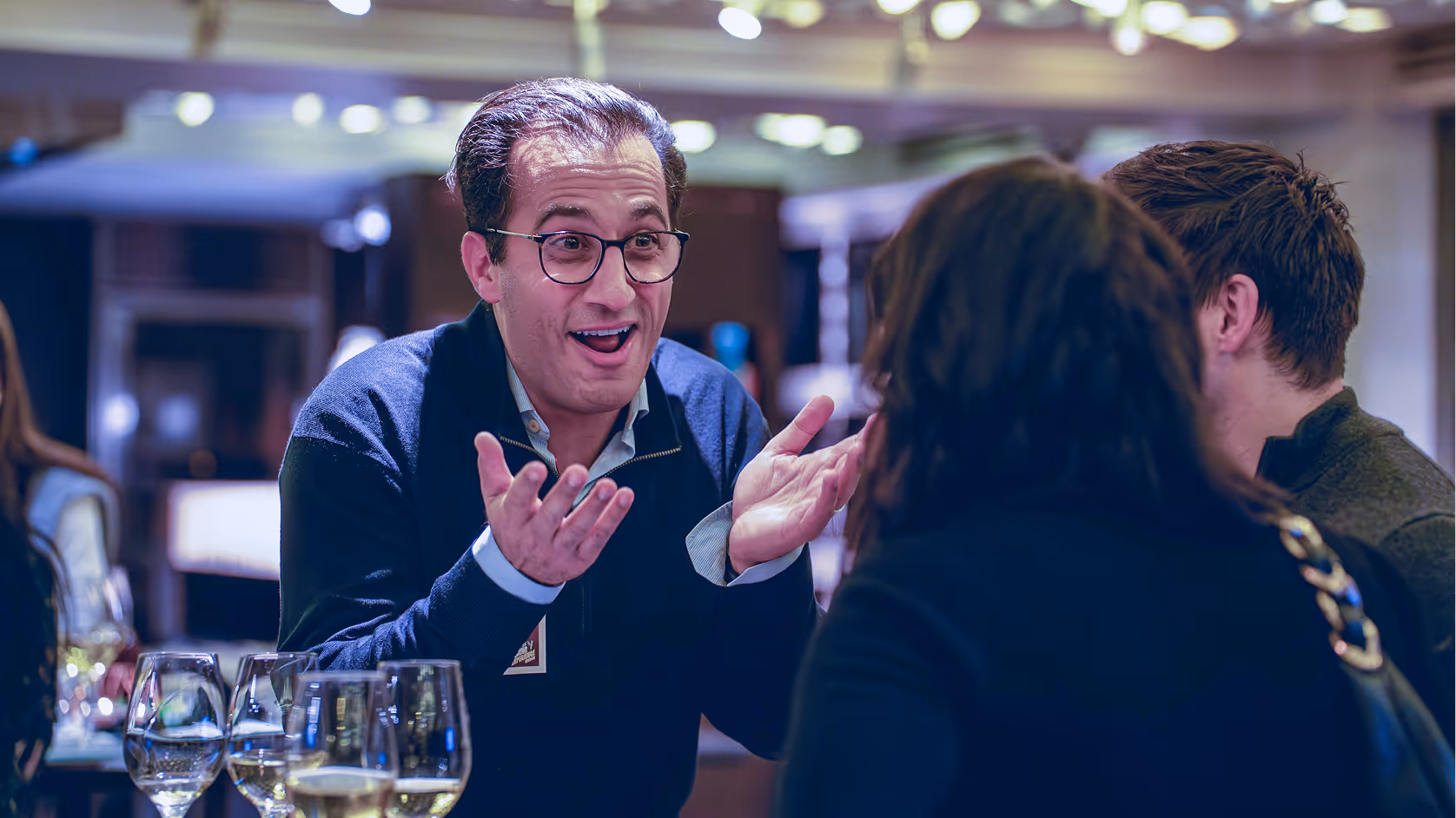 Man wearing glasses animatedly talking to two people at a social gathering with wine glasses on the table.