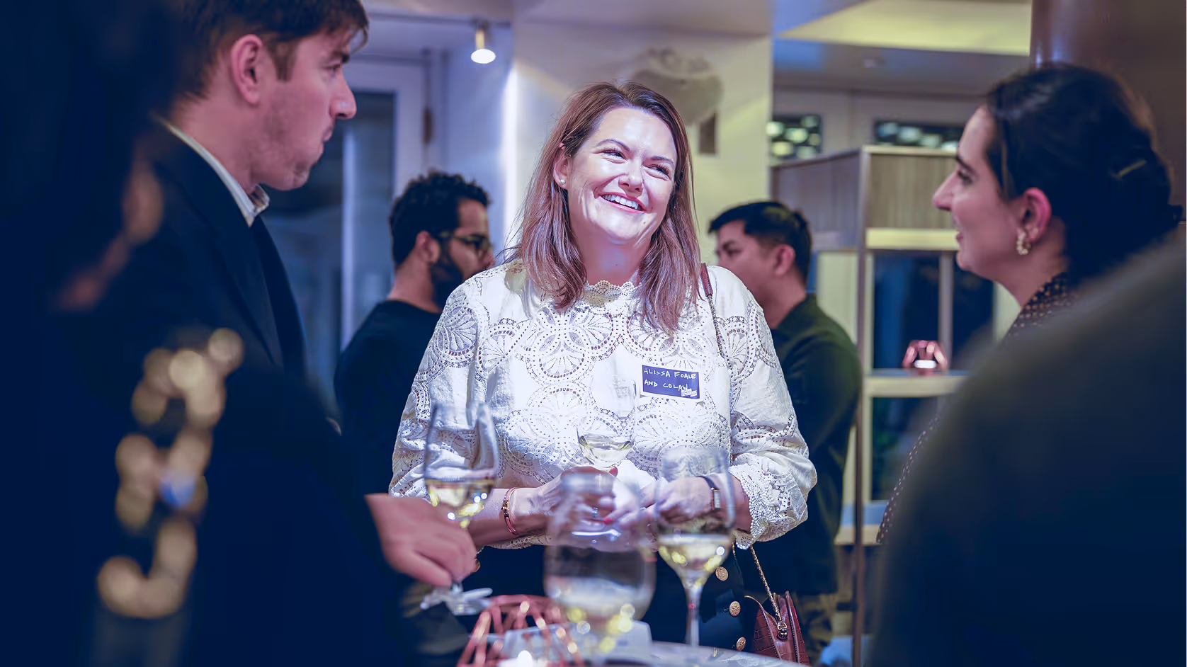 Smiling woman holding a wine glass in a social gathering with people conversing around her.