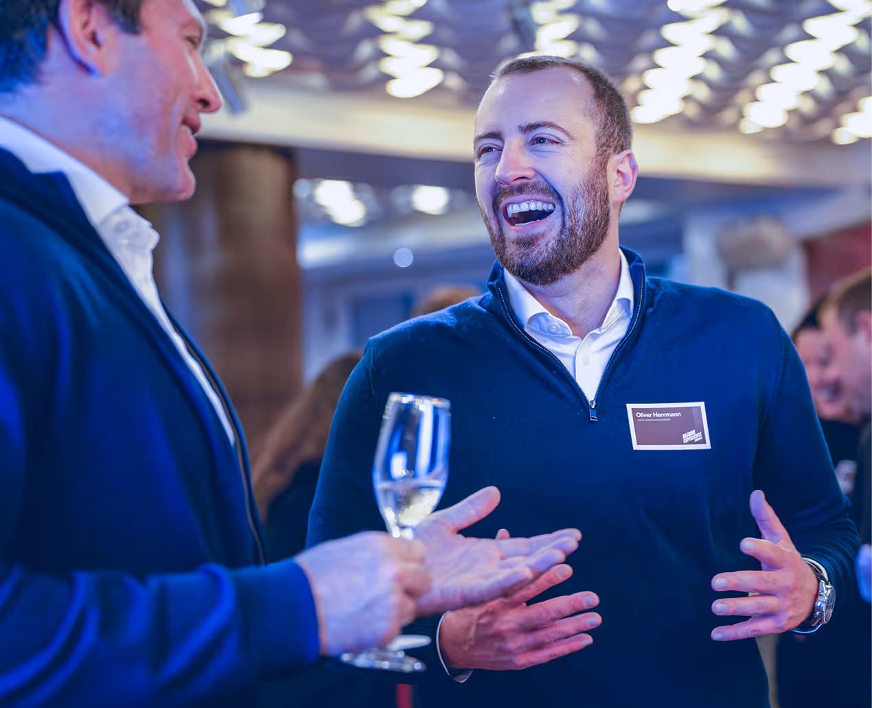 Two men engaged in a lively conversation, one holding a glass of white wine, in a social gathering.