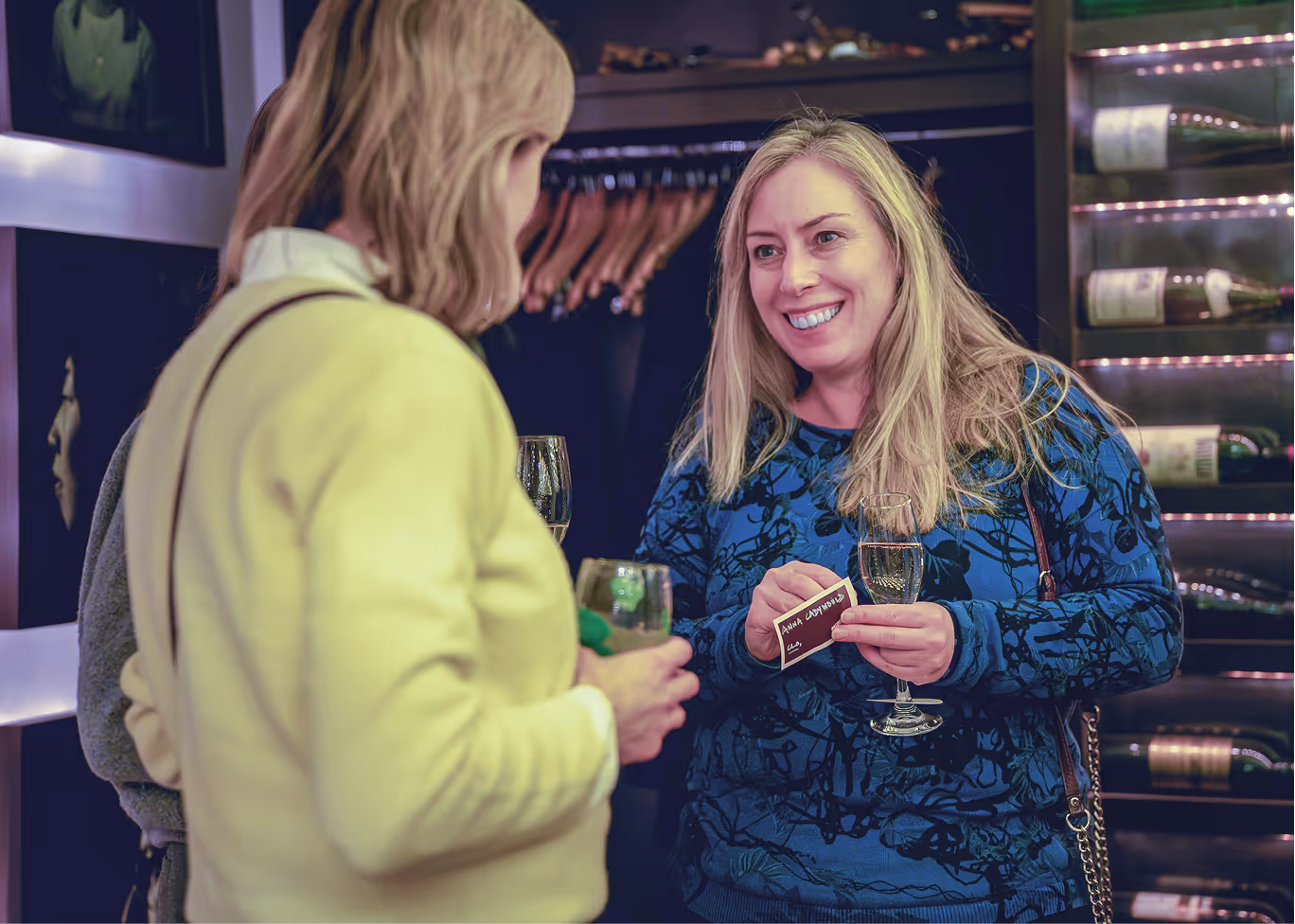 Two women holding wine glasses engaging in conversation, one smiling and holding a name tag.