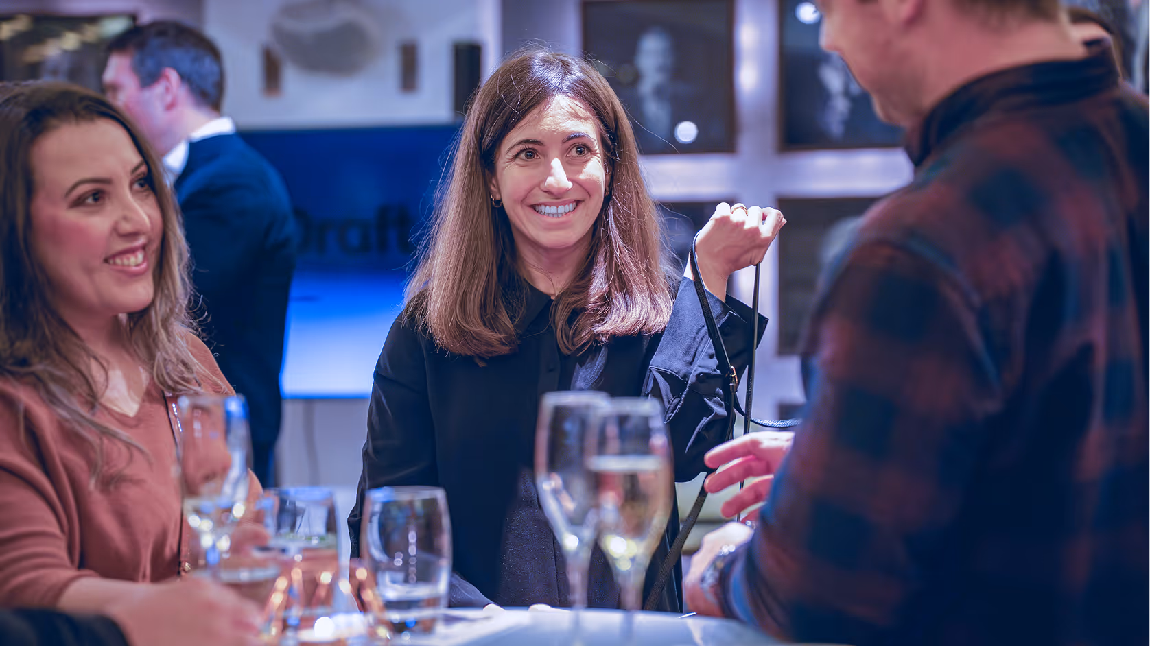 Three people smiling and talking while standing around a table with glasses of drinks.