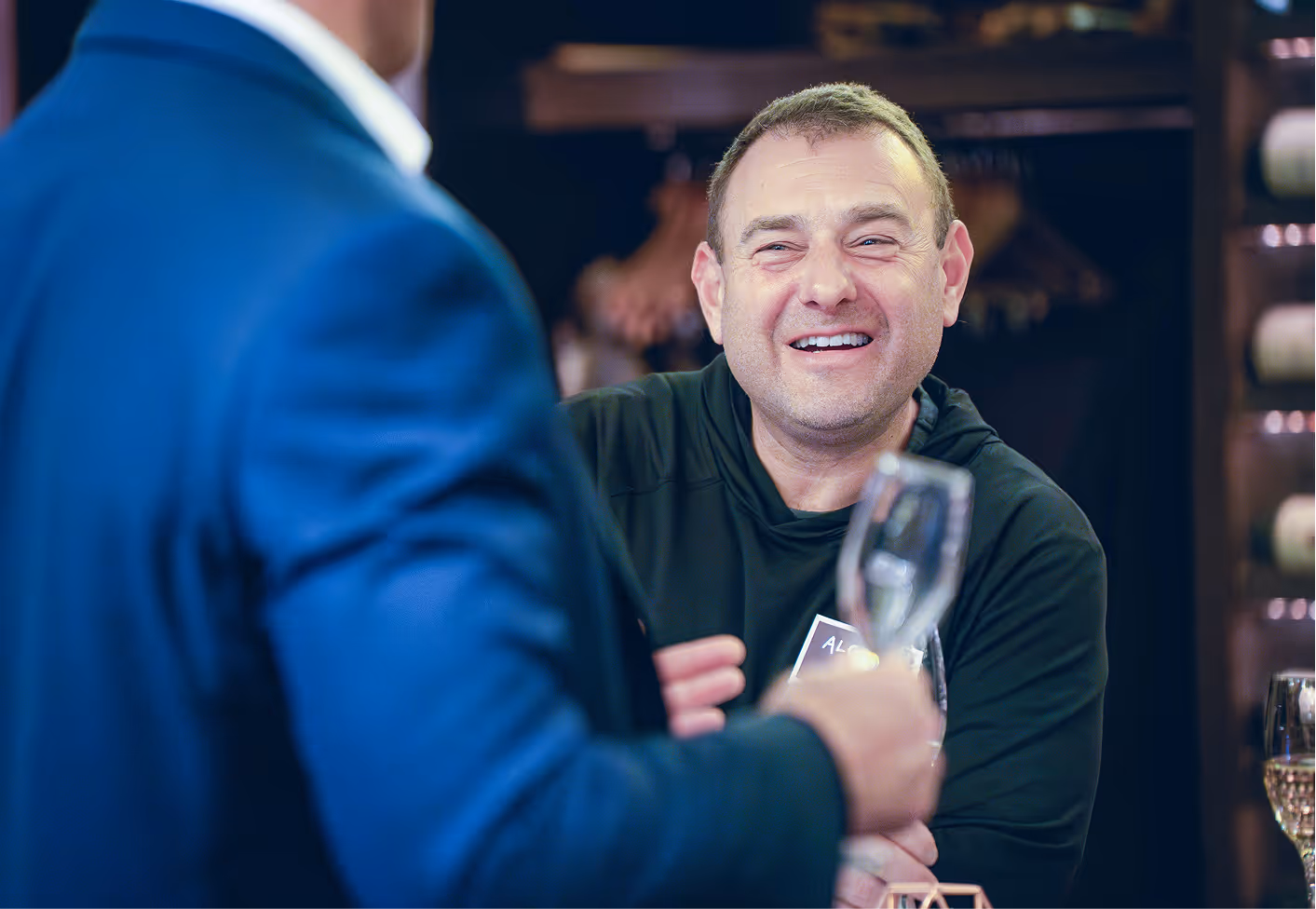 Smiling man in black shirt holding a drink, engaged in conversation with a person in a blue suit.
