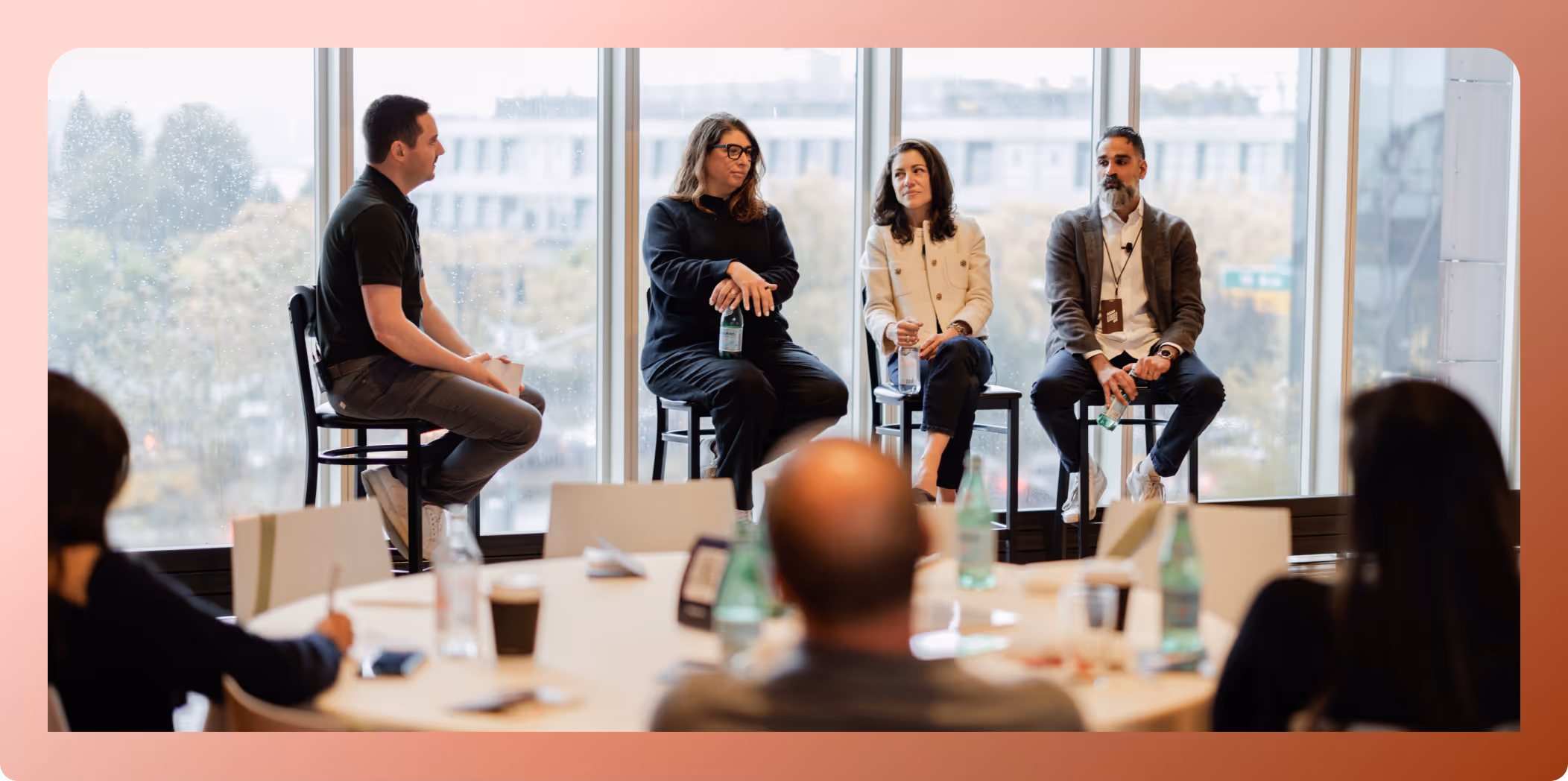 Four people seated on high stools in front of large windows speaking to an audience in a conference room.