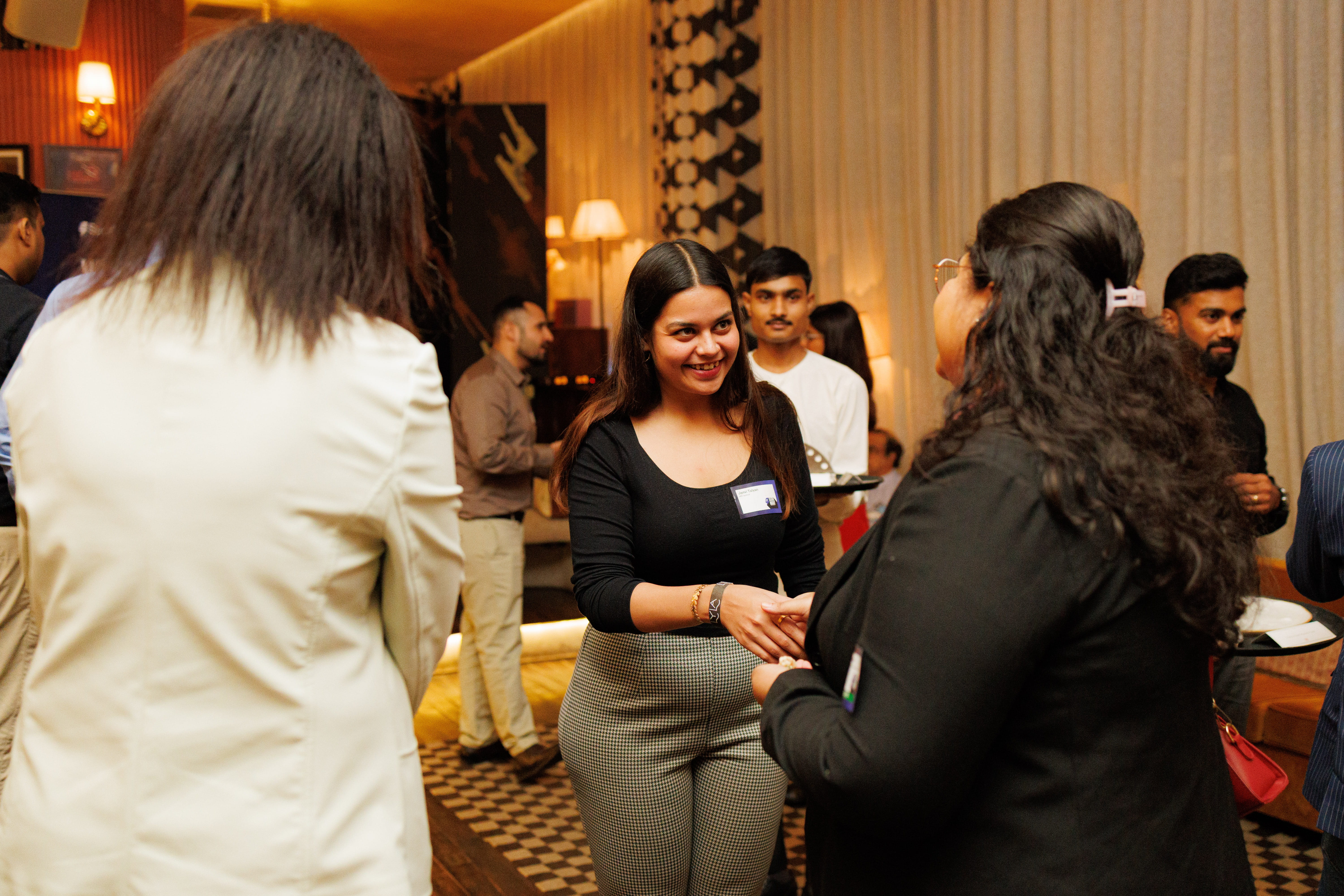 Young woman smiling and shaking hands with another woman at a social indoor event.