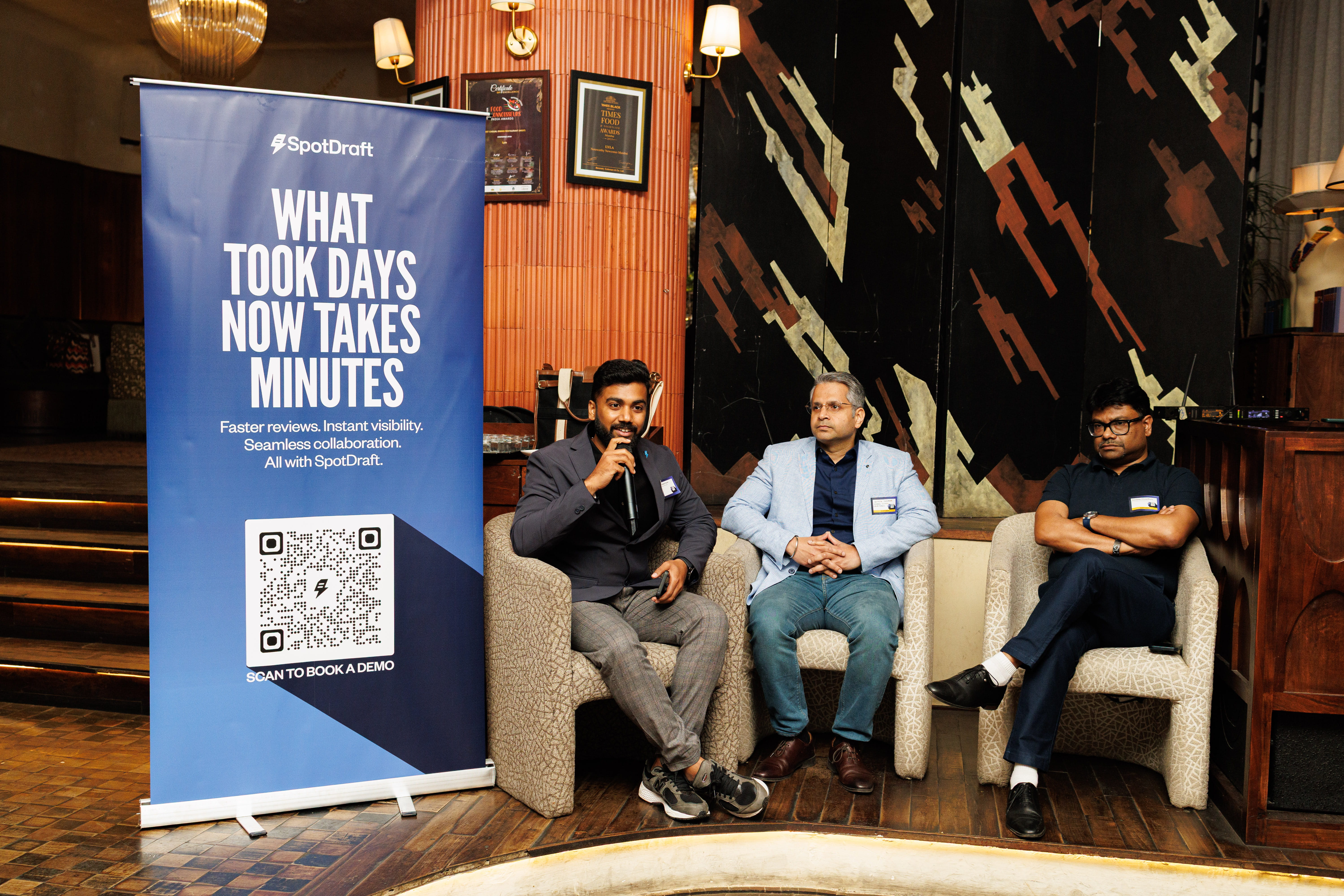 Three men seated on chairs beside a blue SpotDraft banner in a warmly lit room with wooden flooring.