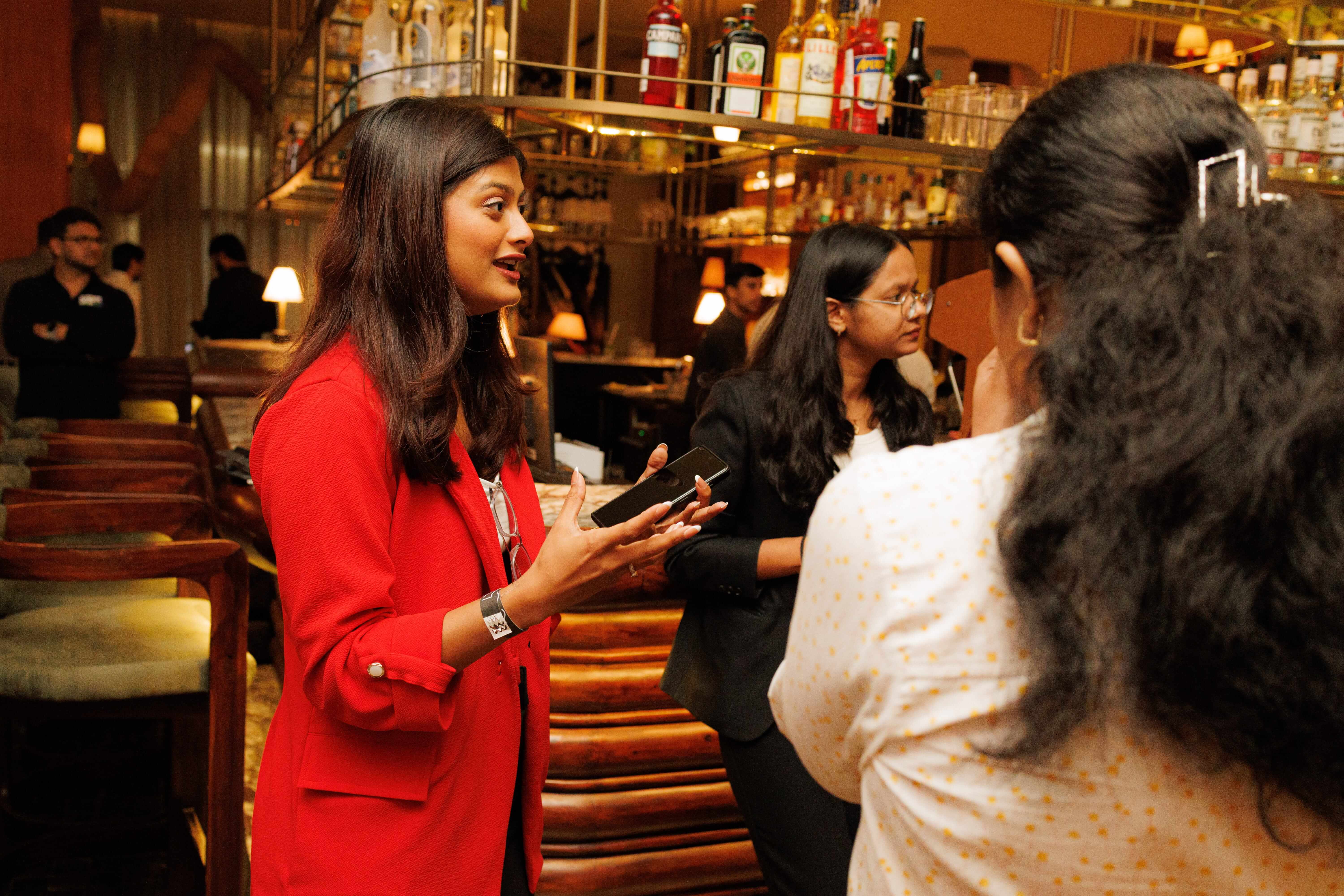 Three women in conversation at a bar, one in a red jacket holding a phone and gesturing.