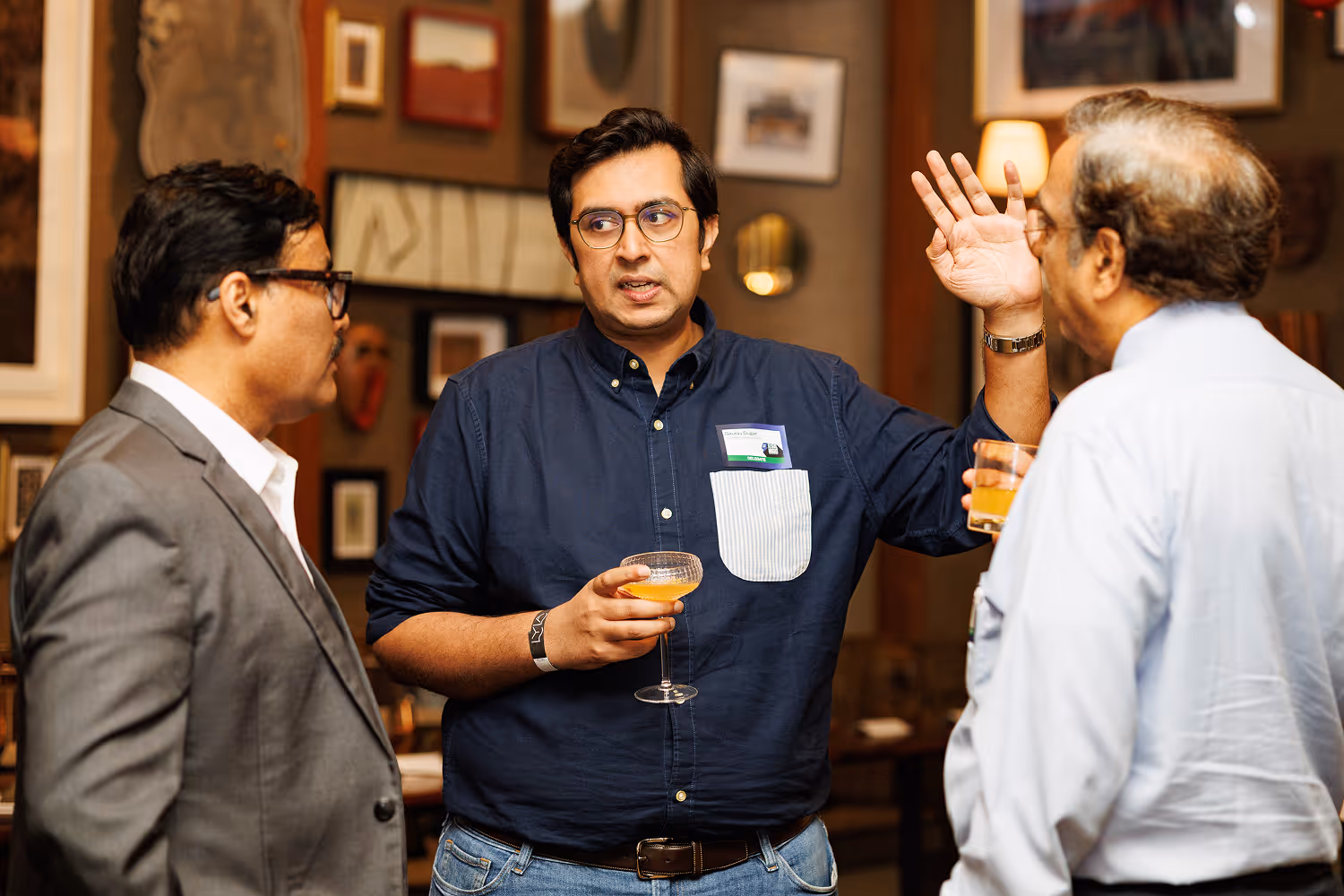 Three men having a conversation indoors, two holding drinks, one gesturing with raised hand.
