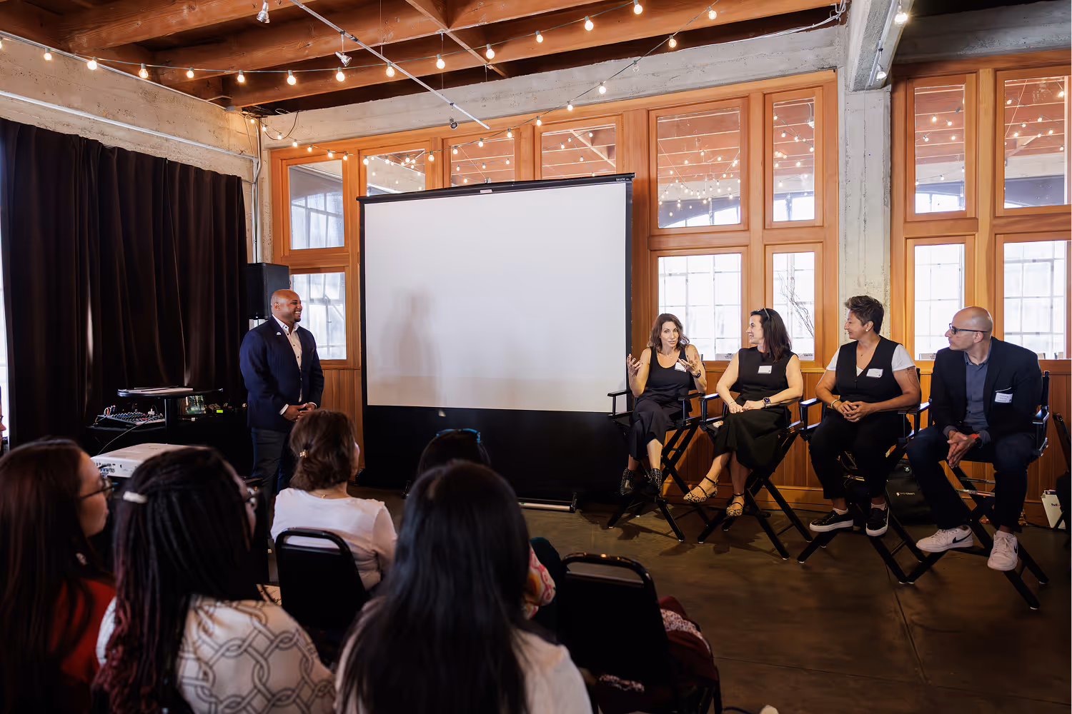 Panel of four people seated with one standing, speaking to an audience in a room with string lights.