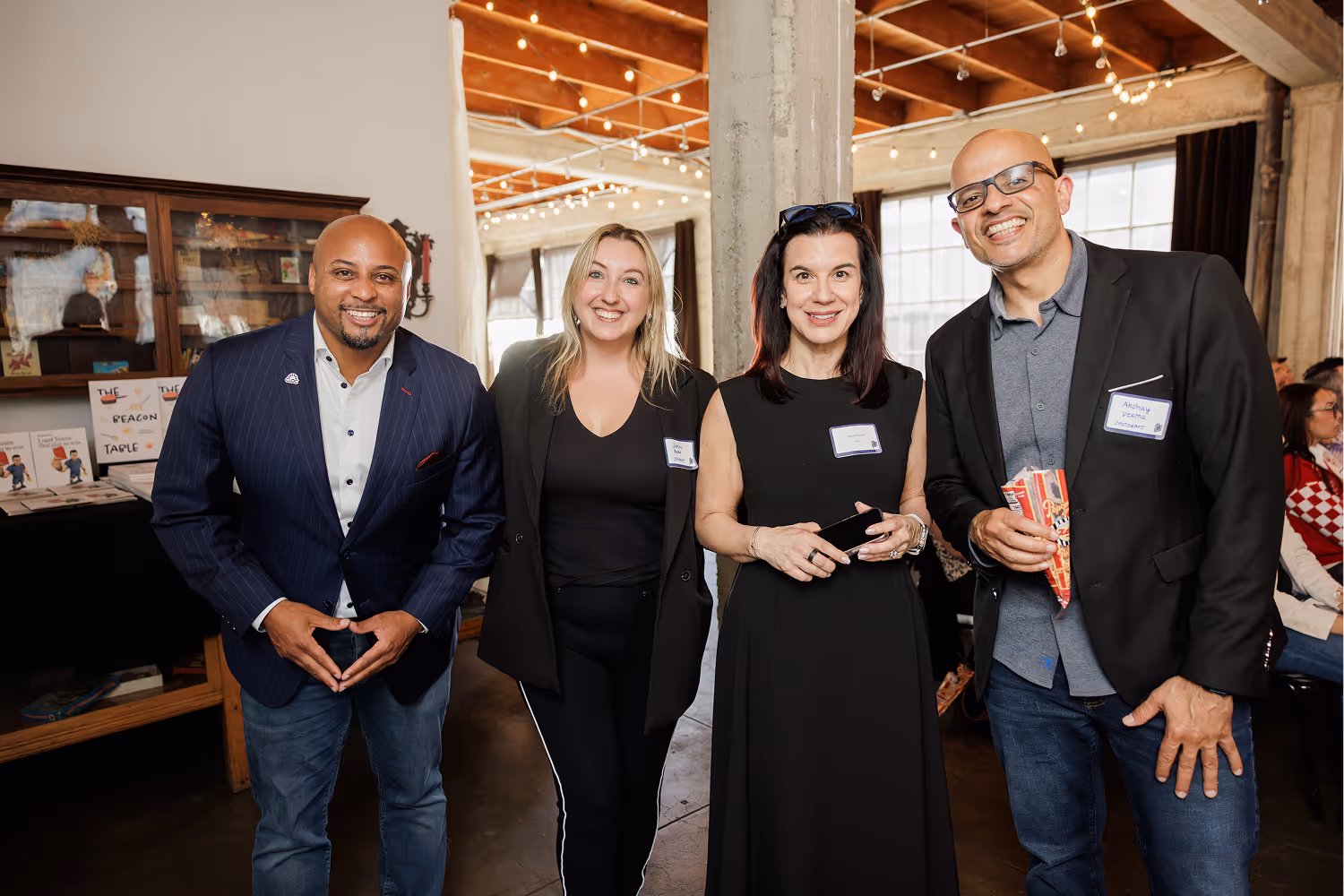 Four smiling people posing indoors at an event, two men and two women wearing name tags and casual business attire.