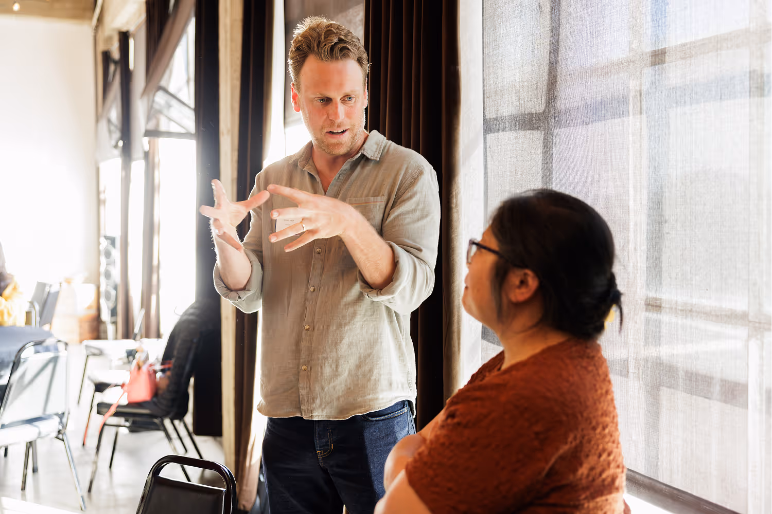 Man with light shirt gesturing and talking to woman with glasses near a window with sheer curtains.