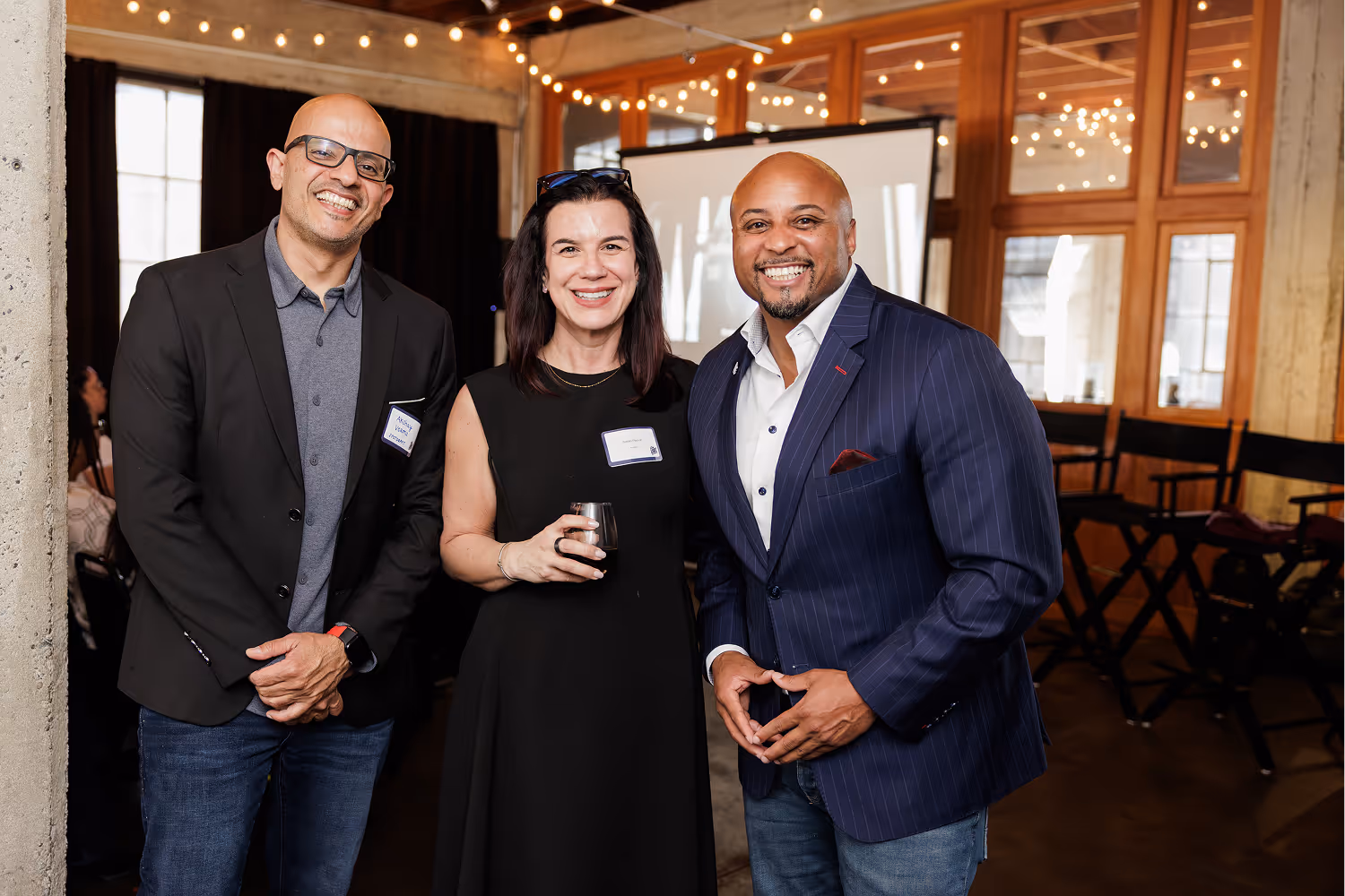 Three adults smiling at a social event, one woman holding a drink between two men in jackets.